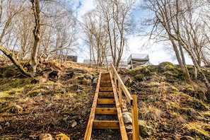 Eine Holztreppe führt einen Berg hinauf, gesäumt von Bäumen und Felsen, mit einem Metallgeländer.