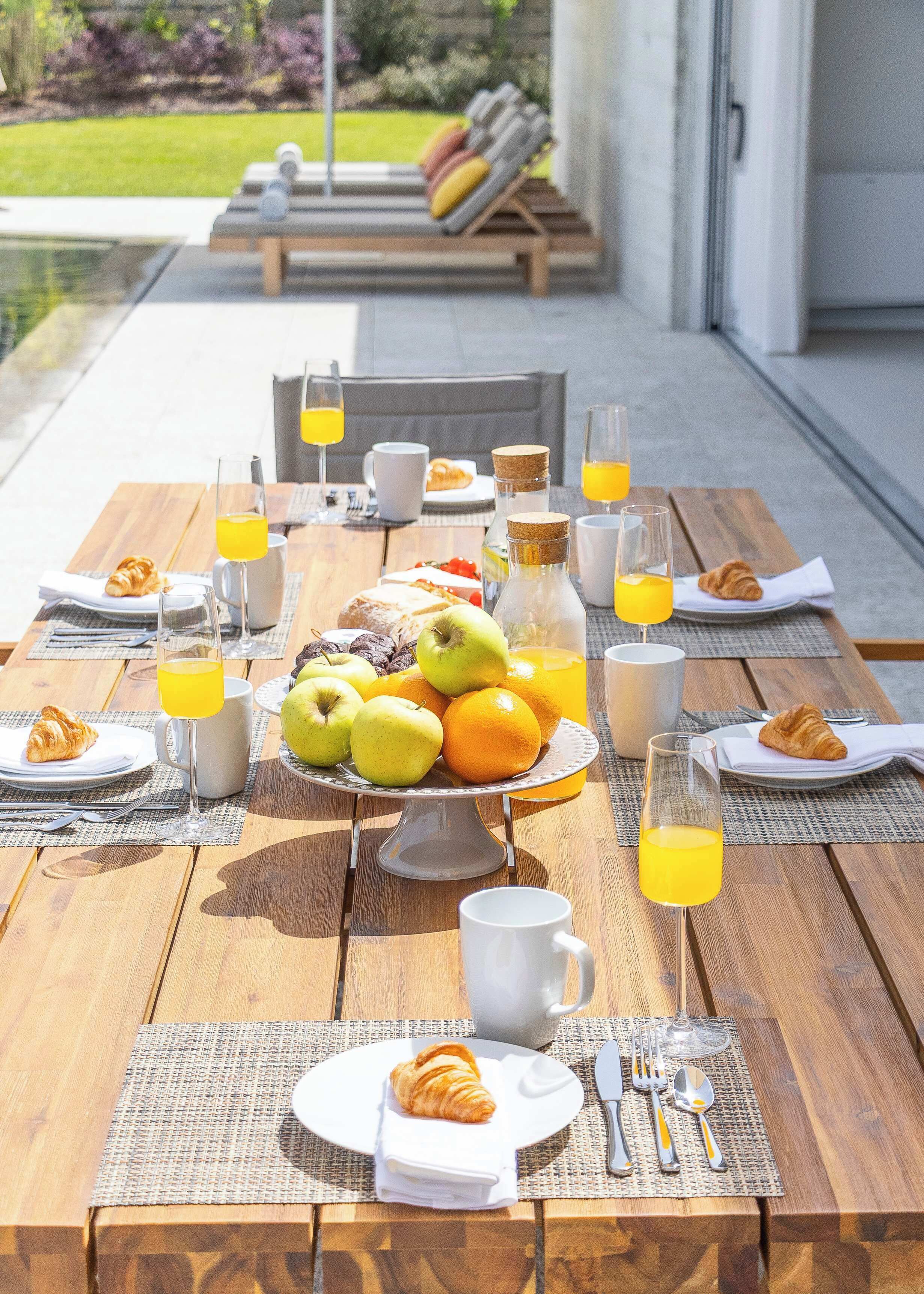 An outdoor breakfast setup with a table, plates, croissants, apples, oranges, and juice glasses.
