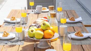 An outdoor breakfast setup with a table, plates, croissants, apples, oranges, and juice glasses.