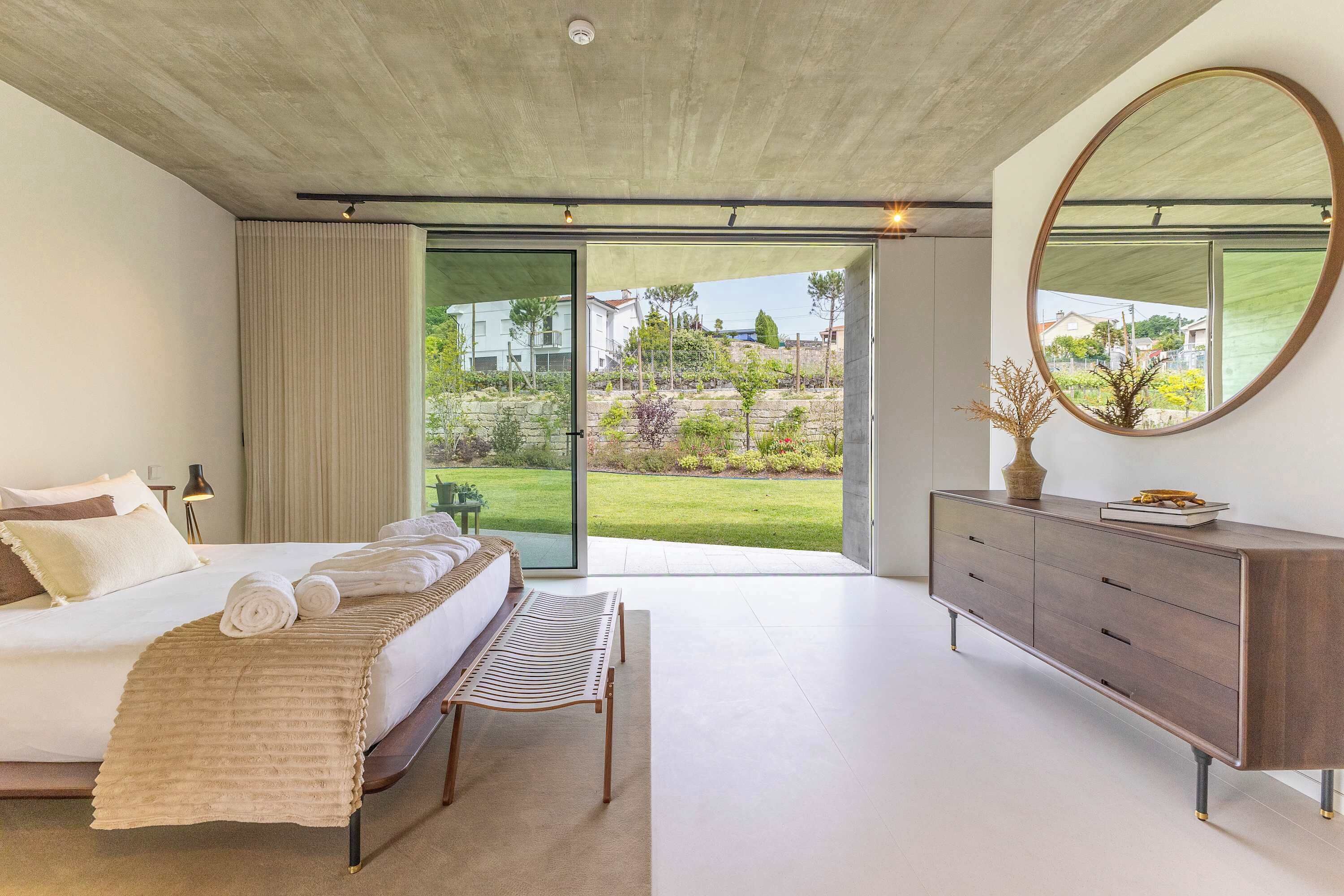 Une chambre à coucher comprenant un lit, un banc, une commode, un miroir et une porte-fenêtre coulissante donnant sur un jardin.