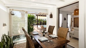 Un coin repas avec une table en bois dressée pour quatre personnes, entourée de chaises en osier, et offrant une vue sur une piscine et un espace de détente extérieur.