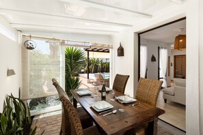 A dining area with a wooden table set for four, surrounded by wicker chairs, with a view of a pool and outdoor seating. - Villa Bananagio - VipVipVillas (Playa Blanca)