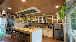 A modern kitchen with a central island, bar stools, wooden ceiling, and potted plants.