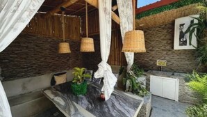A covered outdoor seating area with a stone floor, a table with a marble top, hanging wicker lamps, and a wall with a mural and greenery.