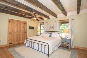 Un dormitorio con cama, mesitas de noche, ventilador de techo, puerta de madera y ventana con cortinas.