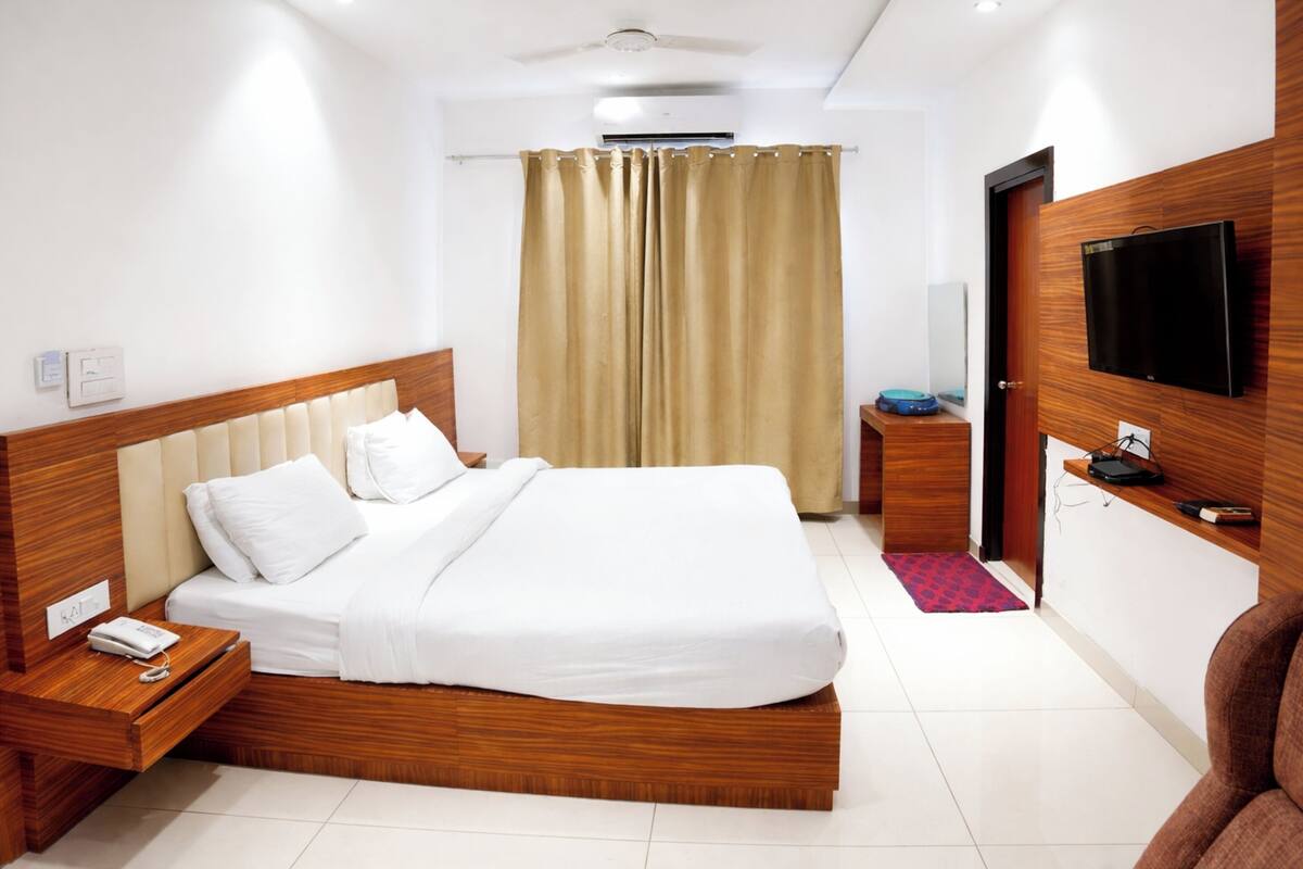 A hotel room with a wooden bed, a TV mounted on the wall, a bedside table with a phone, and a curtain.