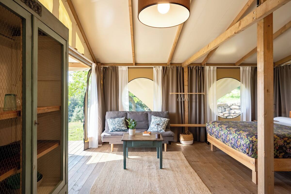 A cozy room with a wooden ceiling, a sofa with patterned cushions, a bed with a floral bedspread, and a small table with a plant.