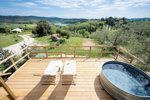 Une terrasse en bois avec deux chaises longues et un jacuzzi, offrant une vue panoramique sur un paysage magnifique, avec un lac et des collines.