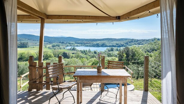 A wooden deck with a table and chairs, a vase with flowers, and a view of a lake and mountains. - Glamping Stregaia (Massa Marittima)