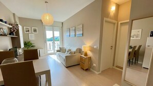 Ein modernes Wohnzimmer mit einer Couch, einem Esstisch und Stühlen. Ein kleiner Beistelltisch mit einer Lampe, eine Deckenleuchte und eine Tür zu einem anderen Raum sind ebenfalls vorhanden.
