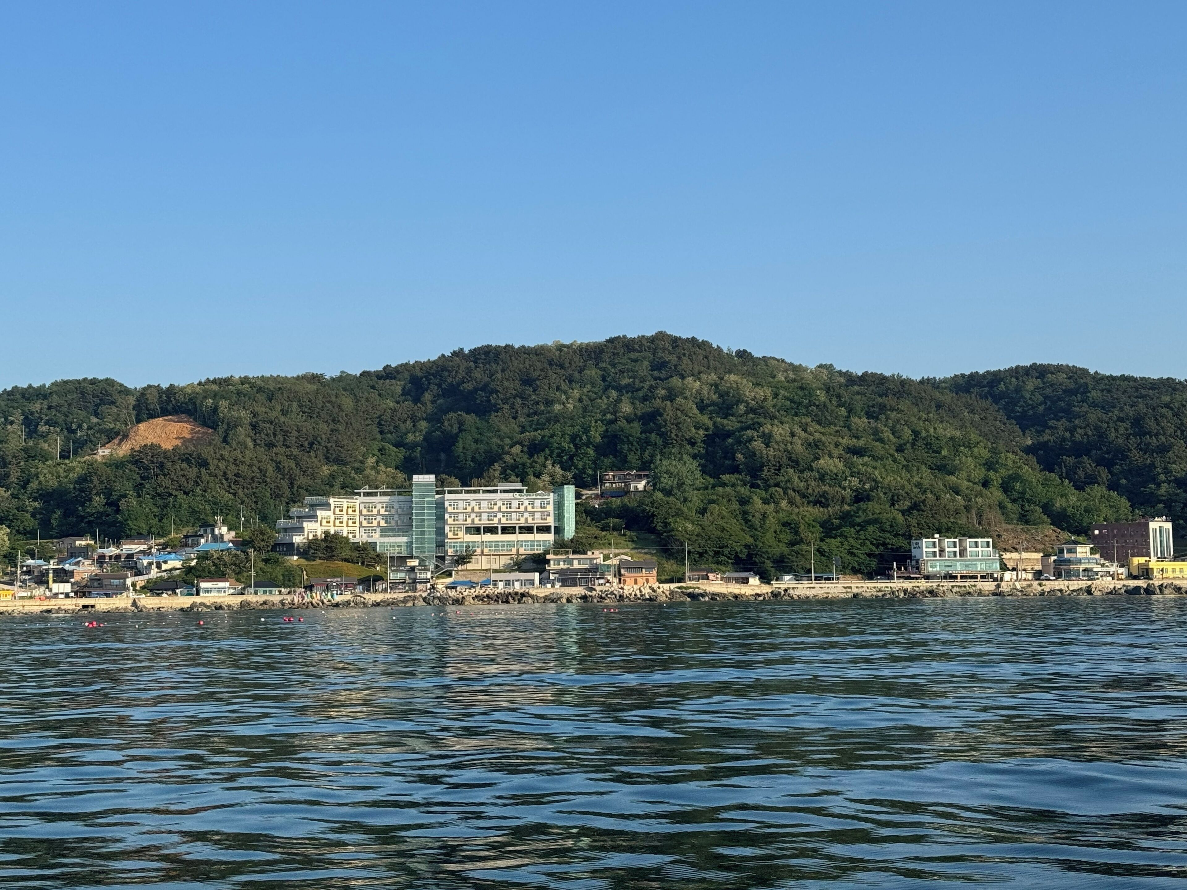 A coastal town with buildings near the water, a hill in the background, and a clear sky.