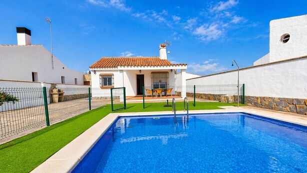 Une piscine avec un espace patio, un bâtiment blanc doté d’un toit en tuiles et un ciel d’un bleu limpide.