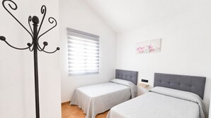 Une chambre avec deux lits, une fenêtre avec des stores, un porte-manteau et une étagère fixée au mur.