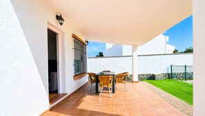 Une terrasse couverte avec une table et des chaises, une fenêtre ornée d’une grille en métal et un mur blanc.
