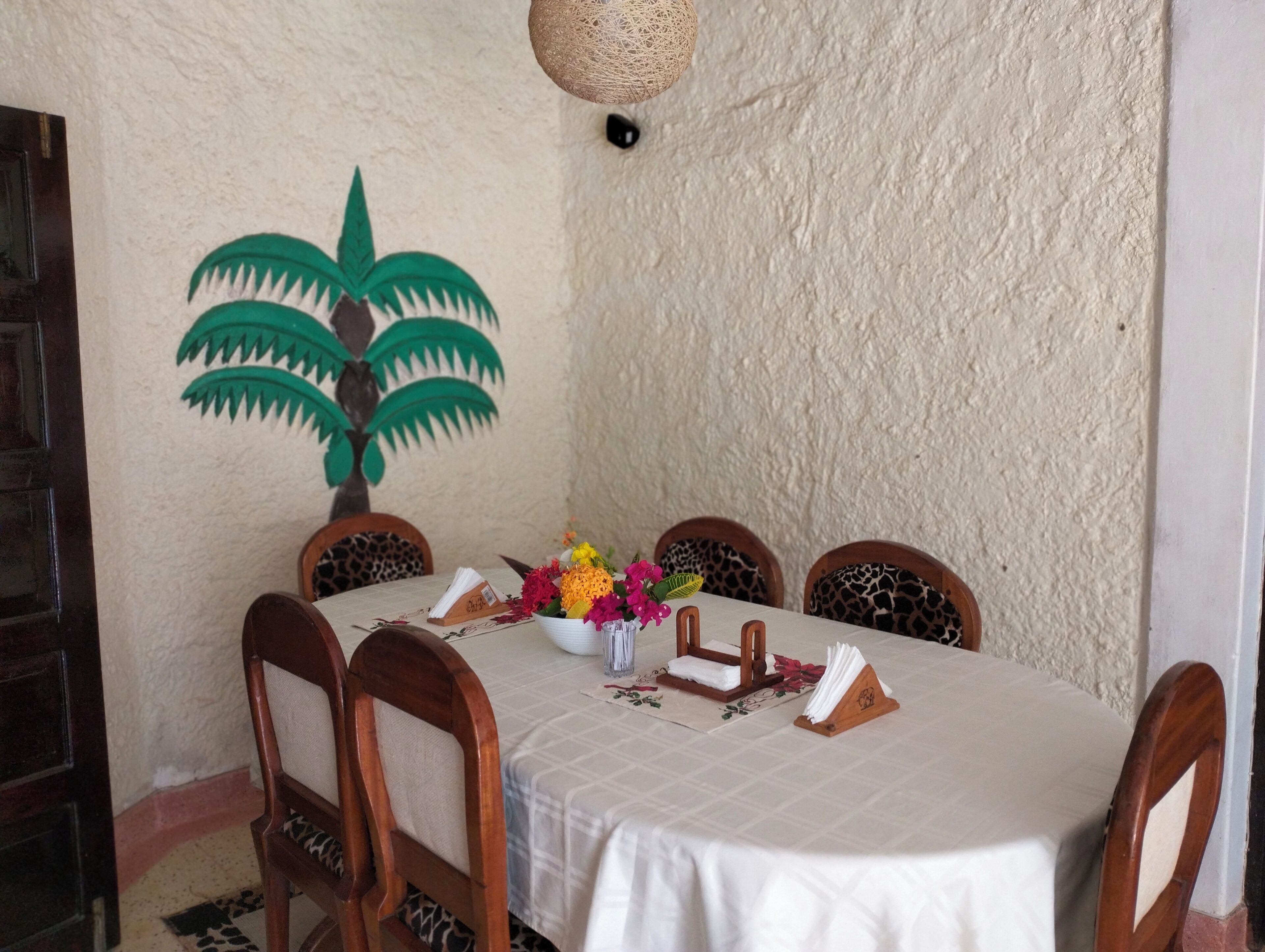 A dining table with a white tablecloth, six wooden chairs with patterned cushions, a woven hanging light fixture, and a wall mural of a palm tree.
