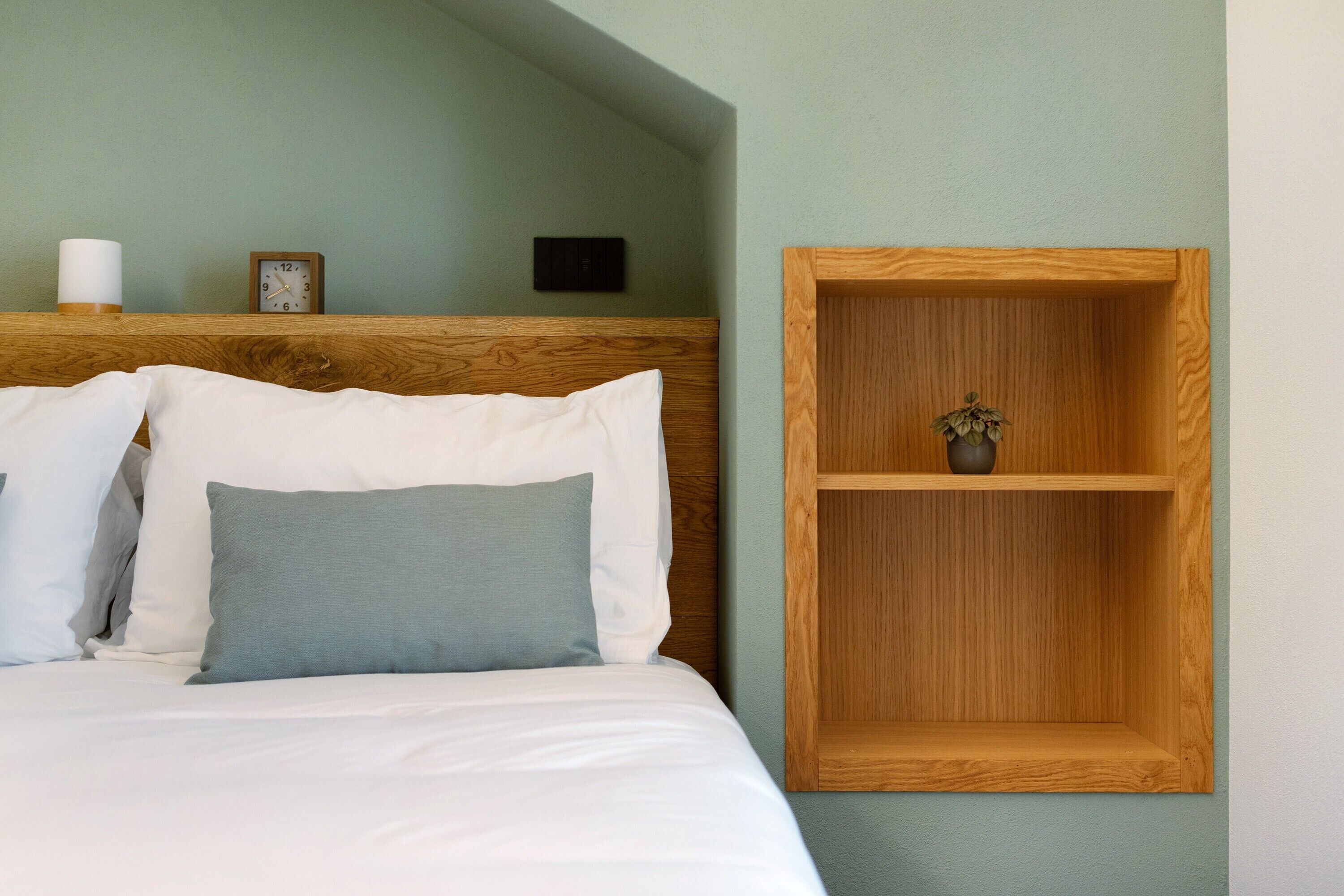 Un letto rifatto con biancheria bianca e grigia, una testiera in legno, un piccolo ripiano in legno con una pianta e una sveglia fissata al muro.