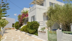 A white building with a stone pathway leading to a gated entrance, surrounded by greenery and a clear sky. - Studios Kymata  (Milos)
