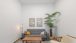 Un salon avec un canapé gris, une table basse en bois, un fauteuil beige, une grande plante en pot et deux tableaux abstraits accrochés au mur.