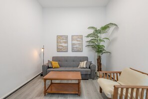 Ein Wohnzimmer mit grauem Sofa, einem Holztisch, einem beigen Sessel, einer großen Topfpflanze und zwei abstrakten Bildern an der Wand.