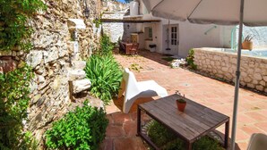 Une cour bordée de murs en pierre, avec une table en bois, des chaises en plastique blanc et un parasol blanc offrant de l’ombre.