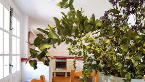 Une table à manger en bois avec des chaises, un vase blanc texturé contenant des feuilles vertes, et une cuisine en arrière-plan.