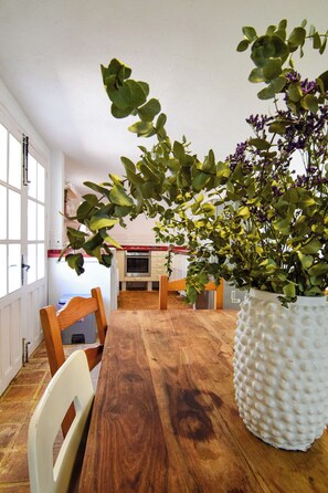 A wooden dining table with chairs, a white textured vase with green foliage, and a kitchen in the background. - La Calma deVilo (Vilo)