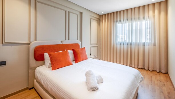 Uma cama bem arrumada com roupa de cama branca e almofadas laranja, piso de madeira e janela com cortinas leves.