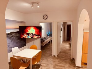 Ein Zimmer mit Fernseher, der ein Feuer zeigt, einem Bett, einem Esstisch mit Stühlen und einer Wanduhr.