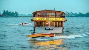 Ein Hausboot mit Deck und überdachter Oberdeck, genannt 'MYTRIP HOUSEBOAT,' auf dem Wasser.
