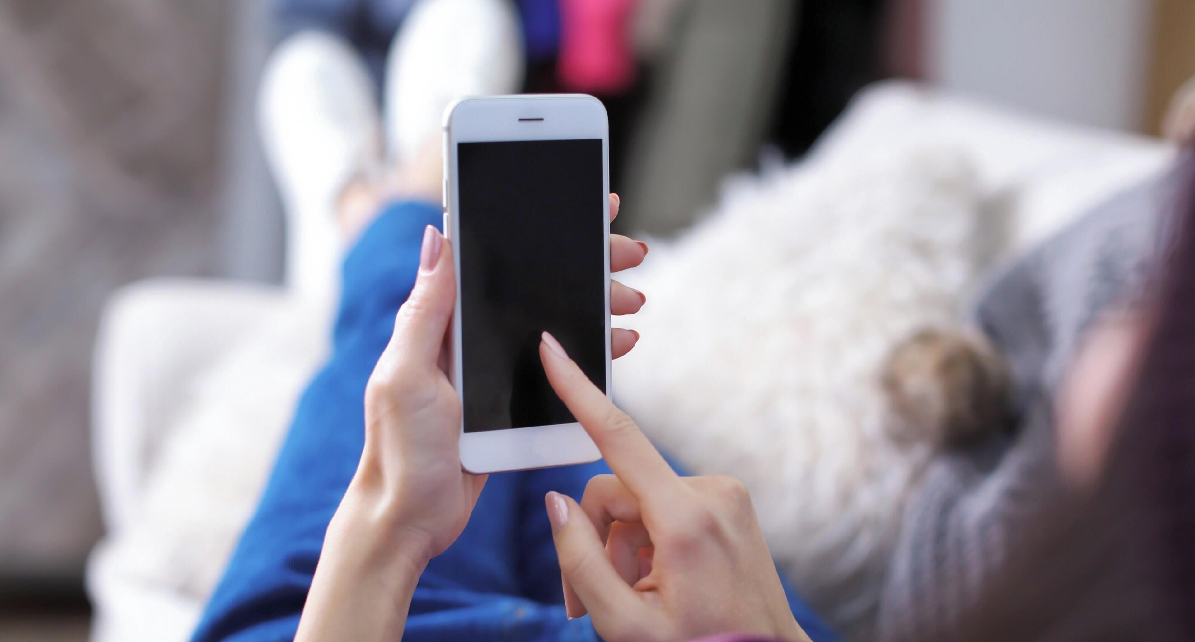 A person holding a smartphone with a black screen, lying on a bed with a white pillow.