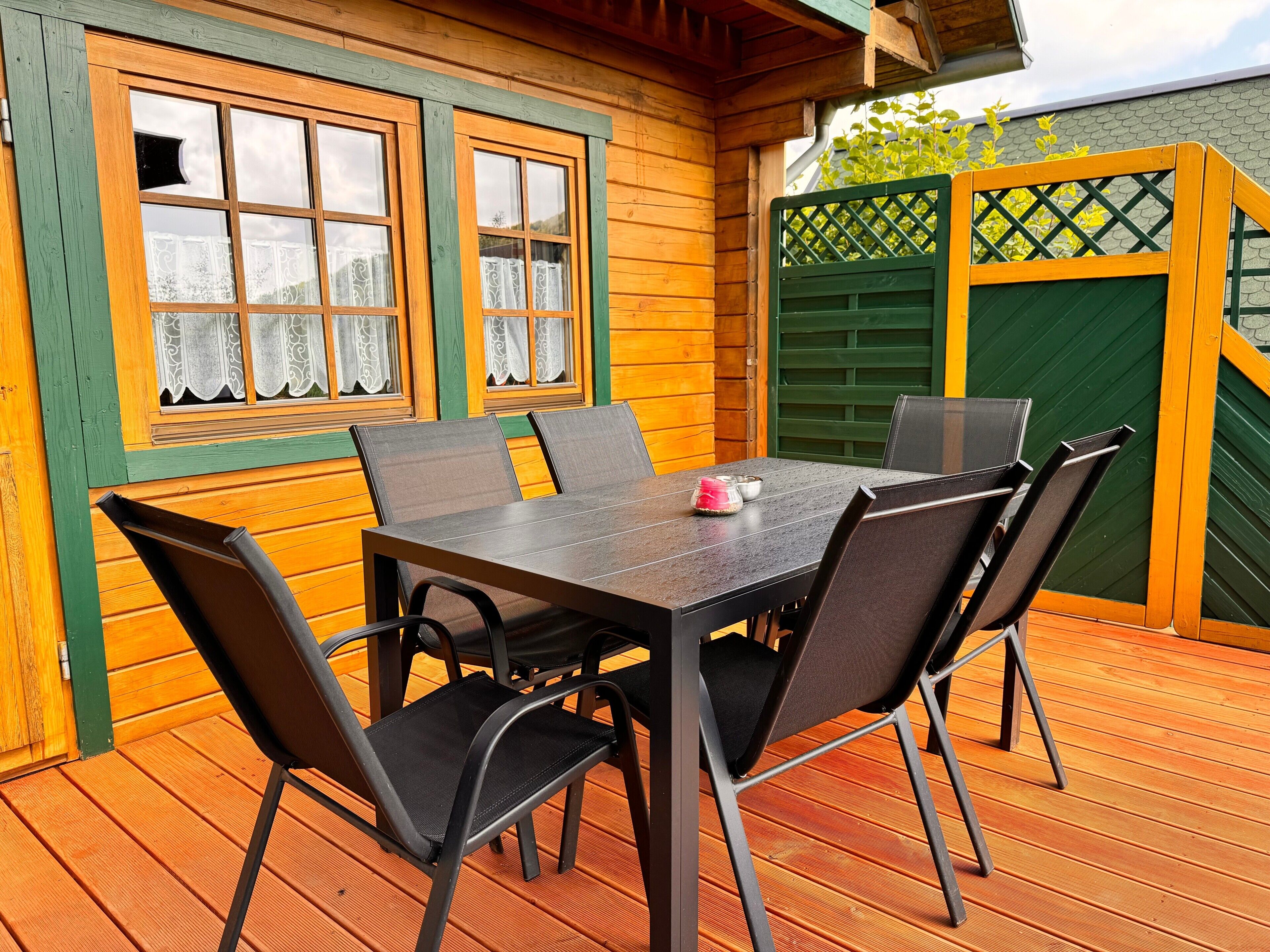 Eine Holzterrasse mit Tisch und Stühlen, ein Fenster mit Spitzenvorhängen und ein grünes Tor.