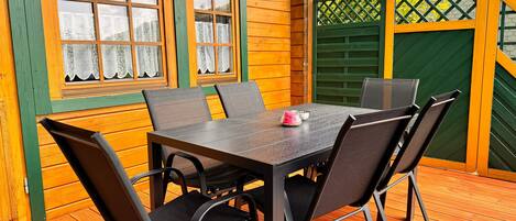 Eine Holzterrasse mit Tisch und Stühlen, ein Fenster mit Spitzenvorhängen und ein grünes Tor.