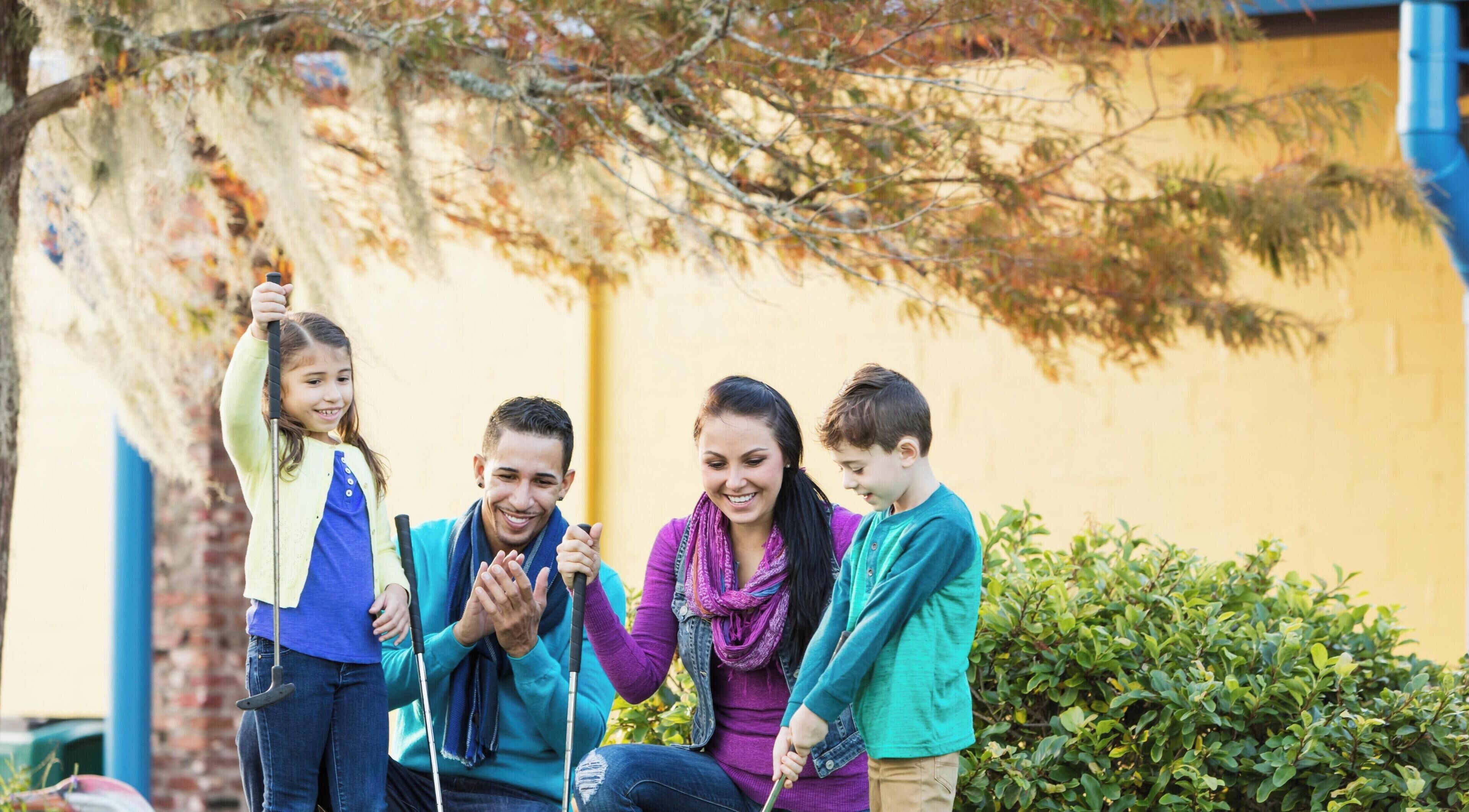A family of four outdoors with a yellow wall and greenery in the background.