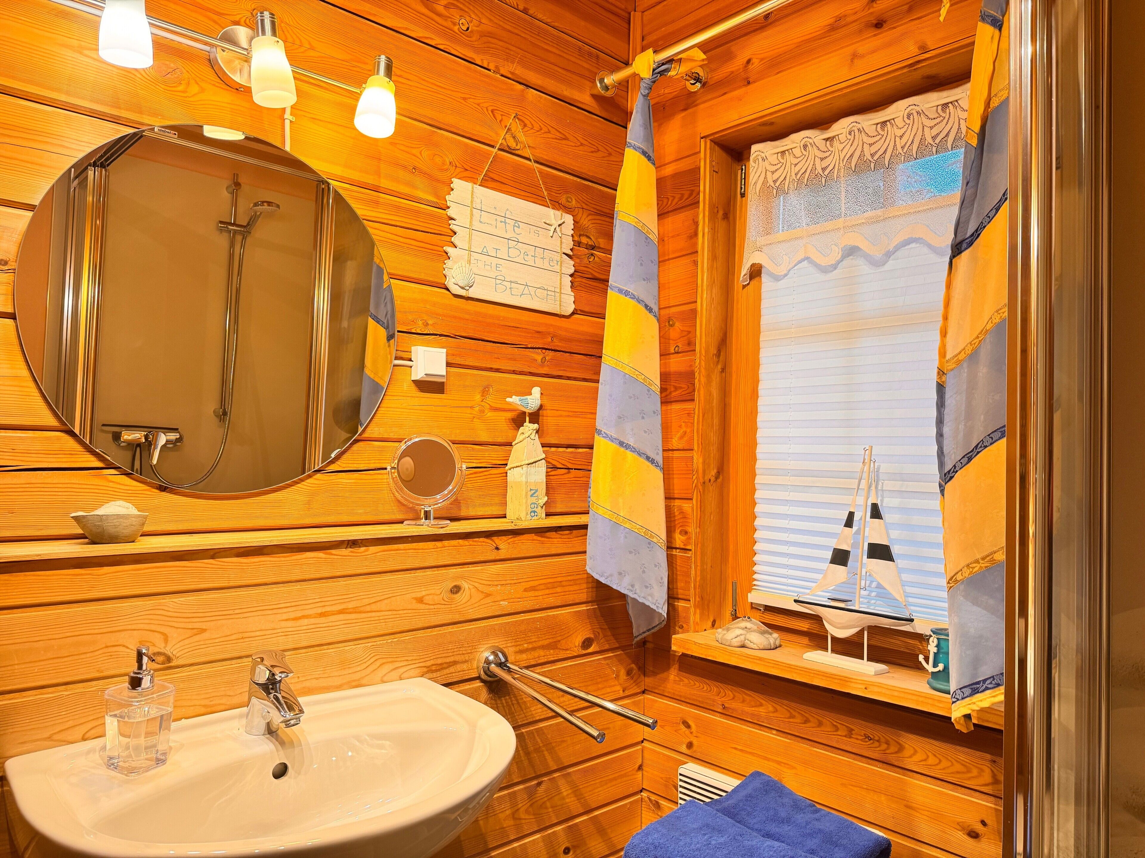 A wooden cabin bathroom with a sink, mirror, towel rack, and a window with a sailboat decoration.