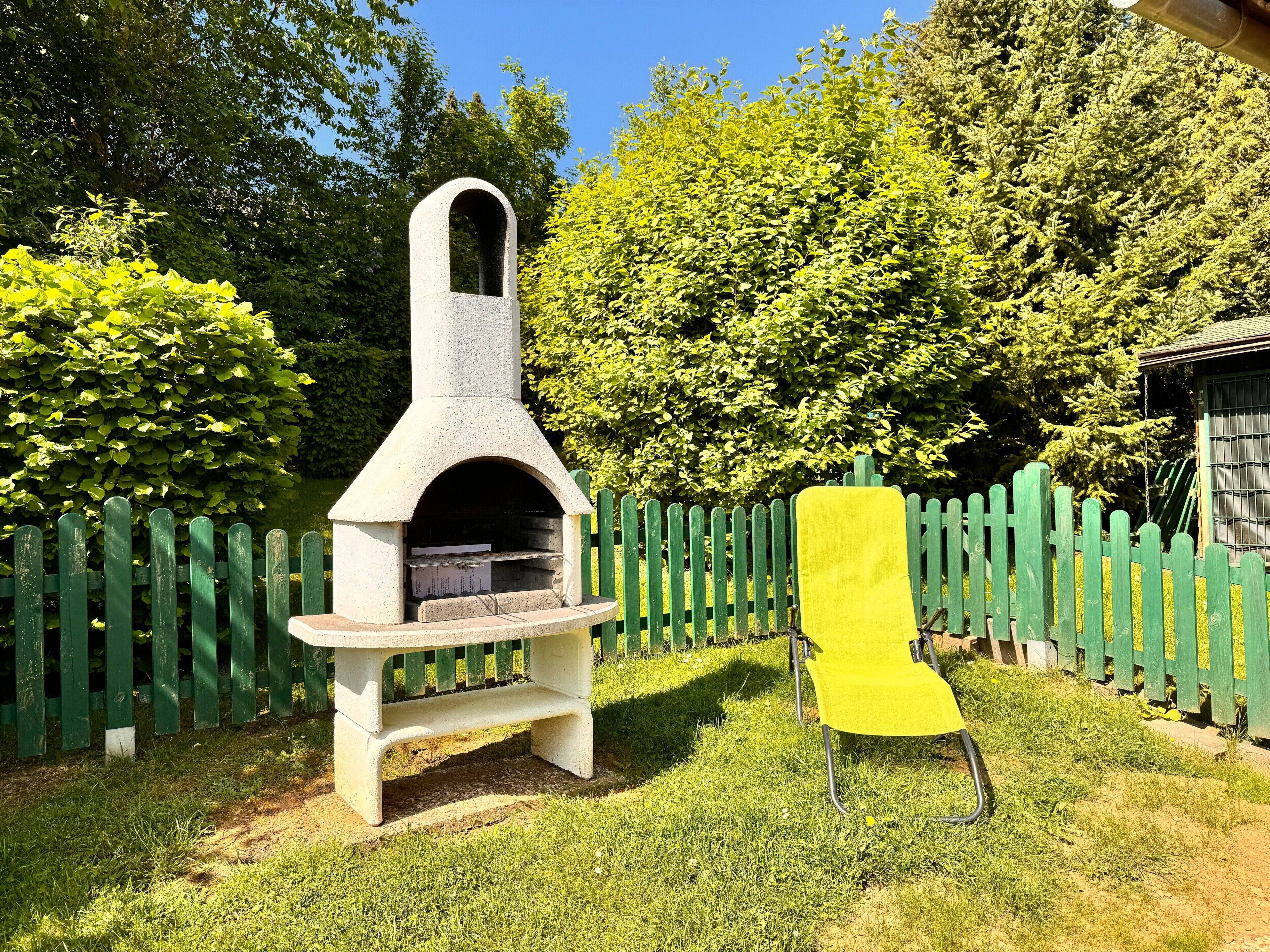 A stone outdoor oven with a bench in a grassy area, surrounded by a wooden fence and trees.