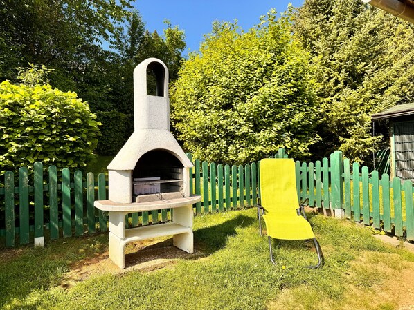 A stone outdoor oven with a bench in a grassy area, surrounded by a wooden fence and trees.