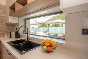 A kitchen with a black sink, a fruit bowl on the counter, and a view of outdoor umbrellas and chairs through a window. - Villa Pietra di Mare-Yeni-Nidri-Lefkada (Geni)