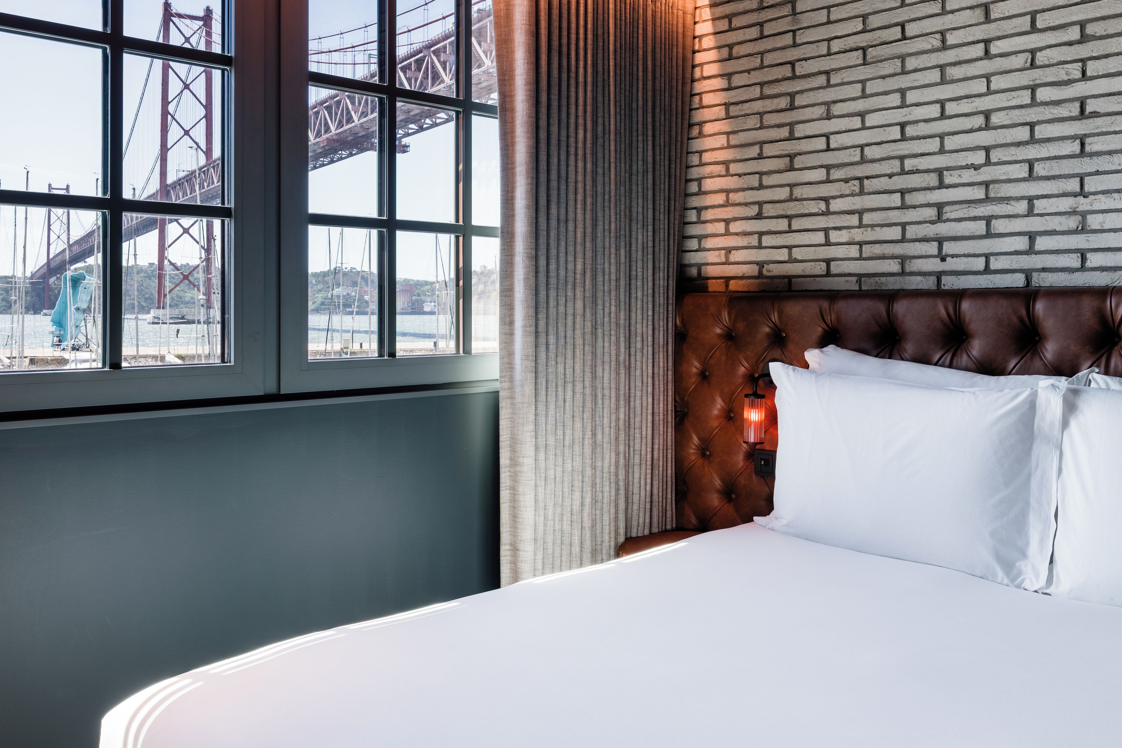 A hotel room with a large window, a brick wall, and a bed with white linens.