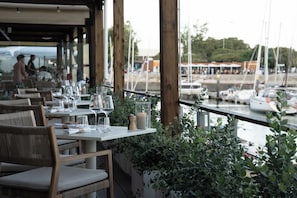 Un área de comedor al aire libre con mesas y sillas, con vista a un puerto deportivo y barcos.