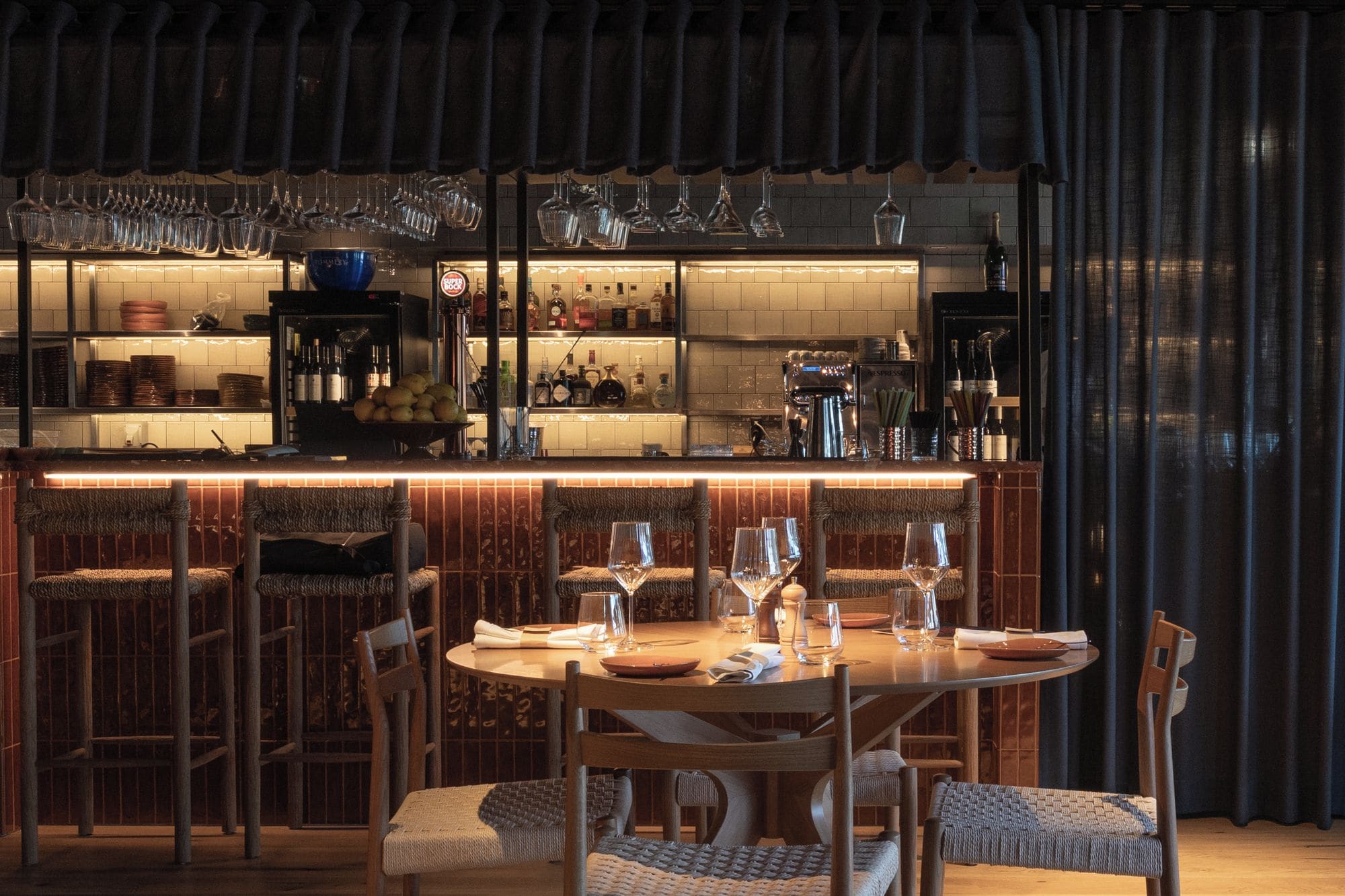 A bar area with a round table and chairs, a variety of bottles and glasses, and a tiled backsplash.