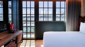 A hotel room with a large window, a bed, a wooden desk with bottles, and a chair.