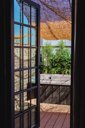 A view through a glass door with a metal grid, leading to an outdoor area with a brick wall, a wooden deck, and green foliage. - Docas Hotel Lisbon (Lisbon)