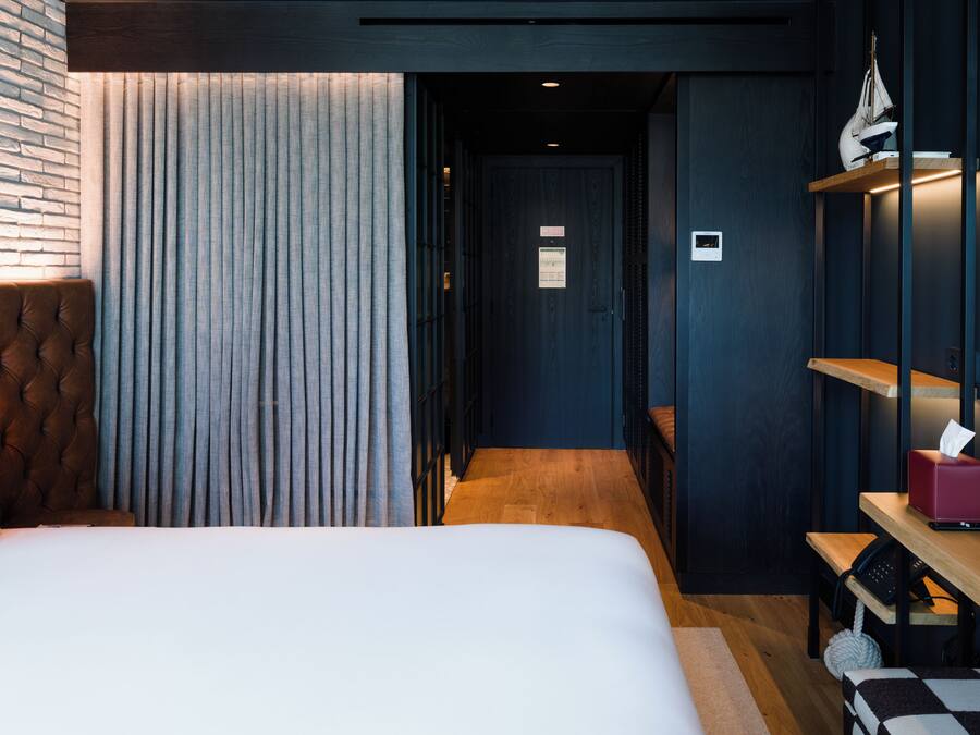 Una habitación de hotel con una cama, una lámpara de noche, un banco de madera, un estante con una caja de pañuelos y una pared con textura.