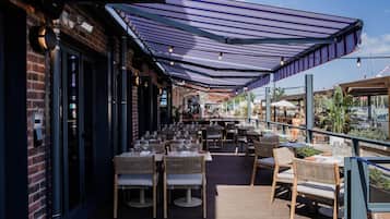 Un área de comedor al aire libre con mesas y sillas, un pasillo cubierto y la fachada de un edificio de ladrillo.