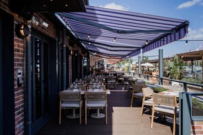 Un espace extérieur aménagé pour les repas, avec des tables et des chaises, une allée couverte et une façade de bâtiment en briques.