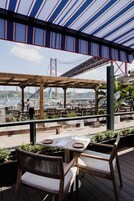 Un área de comedor al aire libre con mesas y sillas, con vista a una marina con barcos y un puente al fondo.