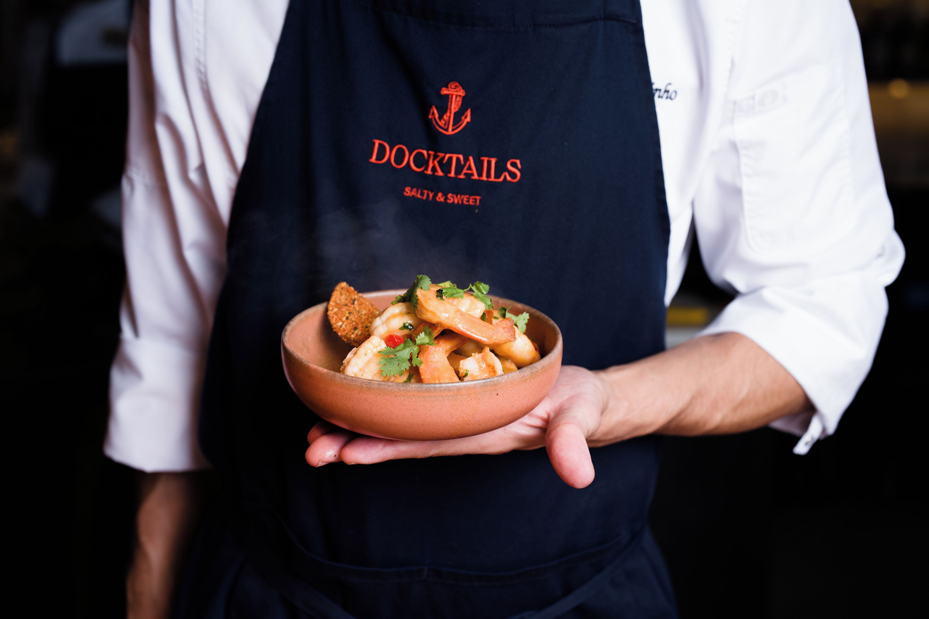 Una persona sosteniendo un plato de comida, con un delantal oscuro que tiene escrito 'DOCKTAILS SALTY & SWEET'.