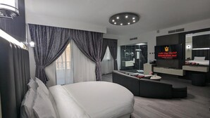 A modern hotel room with a large bed, a seating area, a TV, and a bathroom visible through a glass partition.