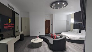 Una camera d'albergo moderna con un divano curvo, un tavolino rotondo, un letto grande e una TV fissata alla parete.