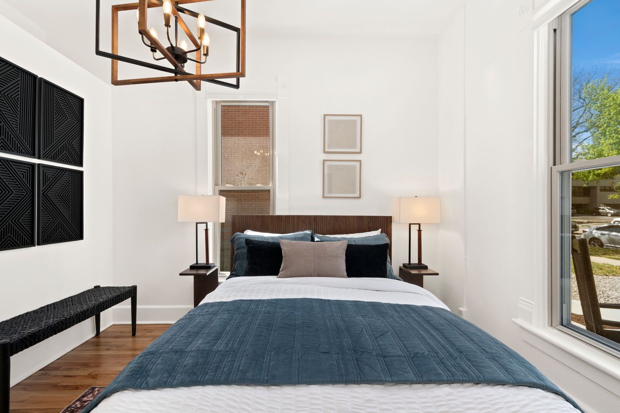 A bedroom with a large bed, two bedside tables with lamps, a bench, a geometric light fixture, a brick wall, and a window with a view.